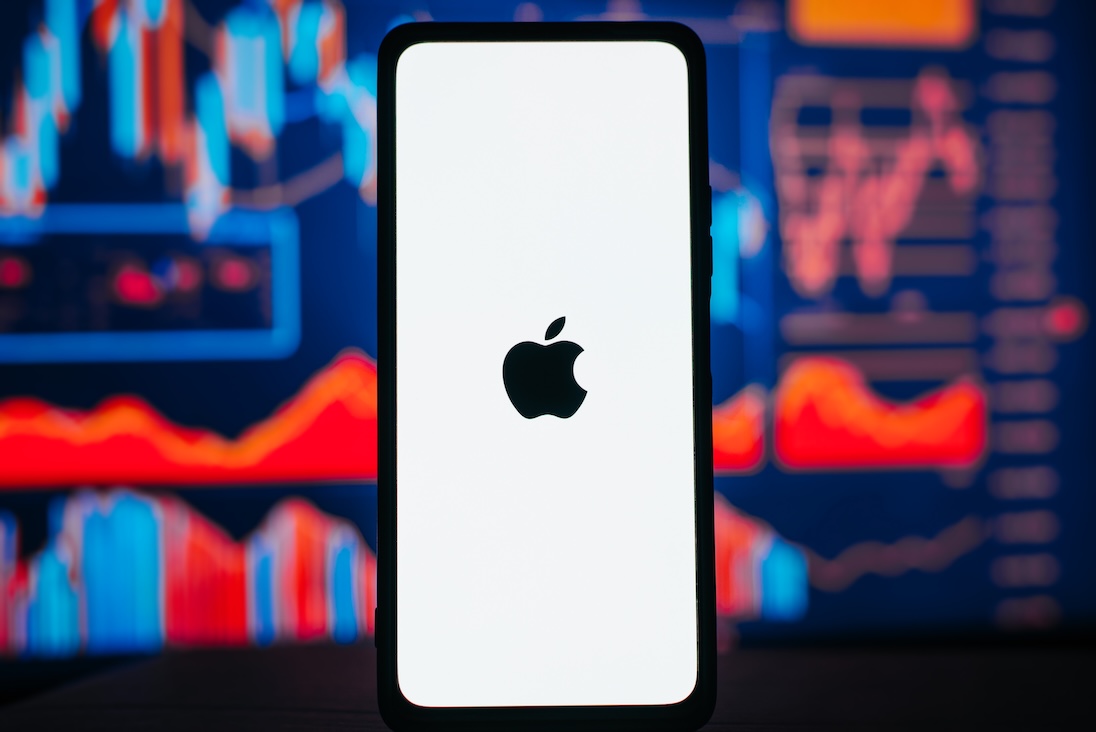 smartphone met logo van APPLE en daarachter een groot scherm met beurskoersen