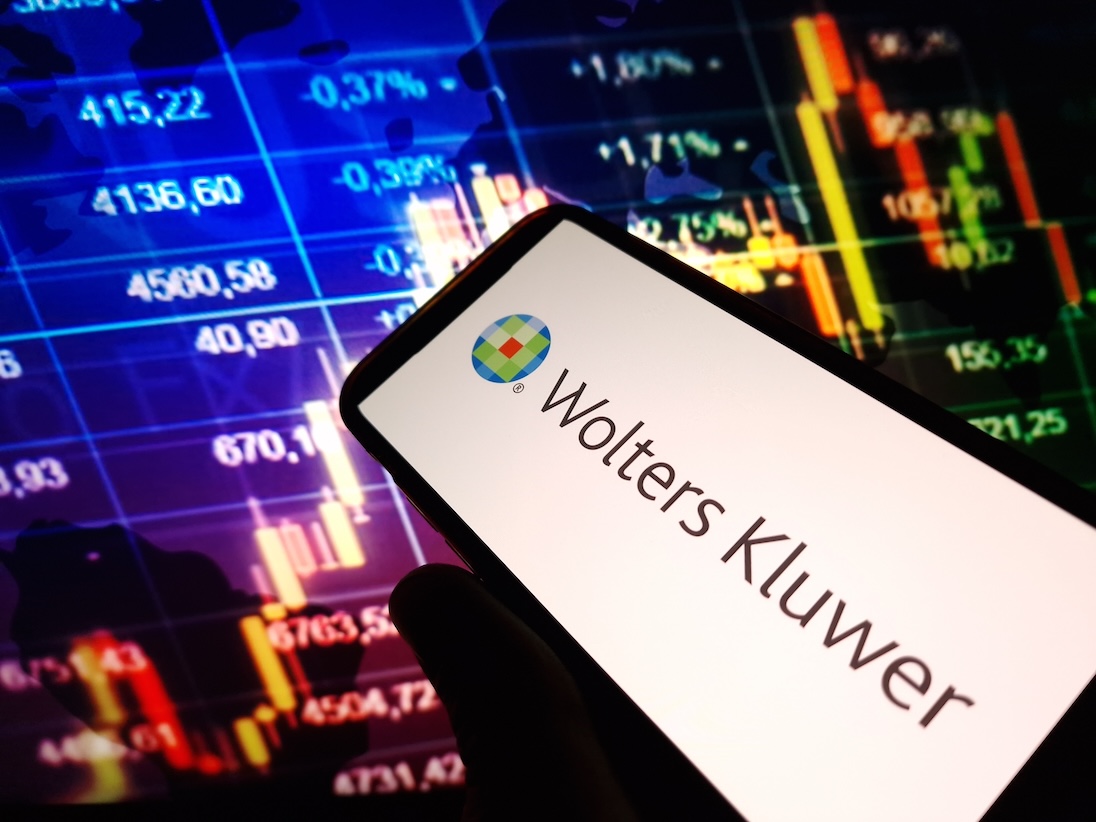 Op scherm van een smartphone letters WOLTERS KLUWER, op achtergrond een scherm met beurskoersen en grafieken